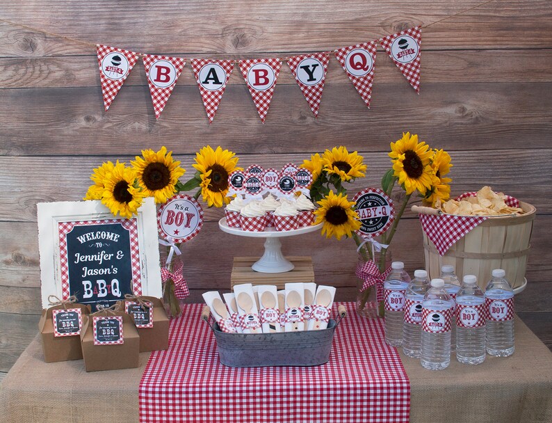 Może przedstawiać: St&oacute;ł na baby shower z czerwono-białym obrusem w kratkę. Na stole znajdują się babeczki, butelki z wodą i przekąski. St&oacute;ł jest ozdobiony słonecznikami i tabliczką z napisem "Witamy na Jennifer & Jason's BBQ".
