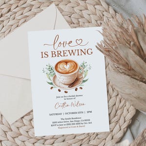 K&ouml;nnte beinhalten: Eine Einladung zur Brautparty mit dem Text "love is brewing" und einem Aquarellbild einer Kaffeetasse mit einem herzf&ouml;rmigen Design im Schaum. Die Einladung befindet sich auf wei&szlig;em Hintergrund mit einem Umschlag und Trockenblumen.