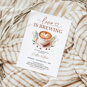 K&ouml;nnte beinhalten: Eine wei&szlig;e Einladungskarte mit dem Text "love is brewing" und einer Aquarellillustration einer Kaffeetasse mit einem herzf&ouml;rmigen Design. Die Karte befindet sich auf einem gestreiften Stoffhintergrund mit getrockneten Pflanzen.