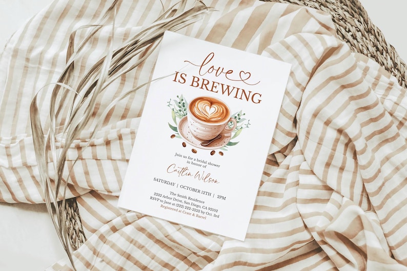 K&ouml;nnte beinhalten: Eine wei&szlig;e Einladungskarte mit dem Text "love is brewing" und einer Aquarellillustration einer Kaffeetasse mit einem herzf&ouml;rmigen Design. Die Karte befindet sich auf einem gestreiften Stoffhintergrund mit getrockneten Pflanzen.