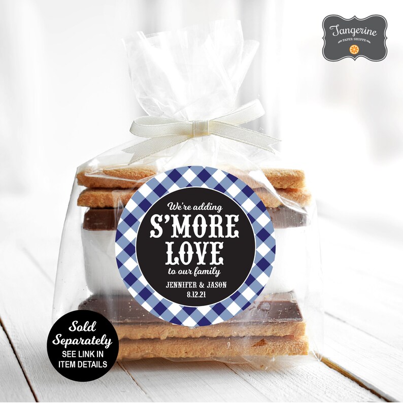 Op de afbeelding: Een ronde sticker met een blauw-wit gingham-patroon met zwarte tekst die luidt "We're adding S'MORE LOVE to our family Jennifer & Jason 8.12.21". De sticker zit op een doorzichtige plastic zak gevuld met graham crackers, chocolade en marshmallows.