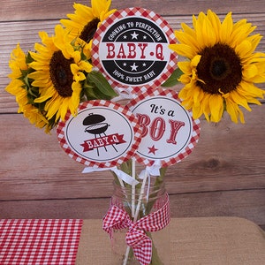 Może przedstawiać: Centralny element dekoracyjny na baby shower z motywem grillowania. Trzy okrągłe papierowe tabliczki z czerwono-białym wzorem w kratkę. Tabliczki przedstawiają napisy "Cooking to Perfection Baby-Q 100% Sweet Baby", "Baby-Q" z obrazkiem grilla oraz "It's a Boy" z czerwoną gwiazdą. Tabliczki są przymocowane do drewnianych szaszłyk&oacute;w i umieszczone w przezroczystym szklanym słoiku z czerwoną i białą wstążką w kratkę. Słoik jest otoczony słonecznikami.