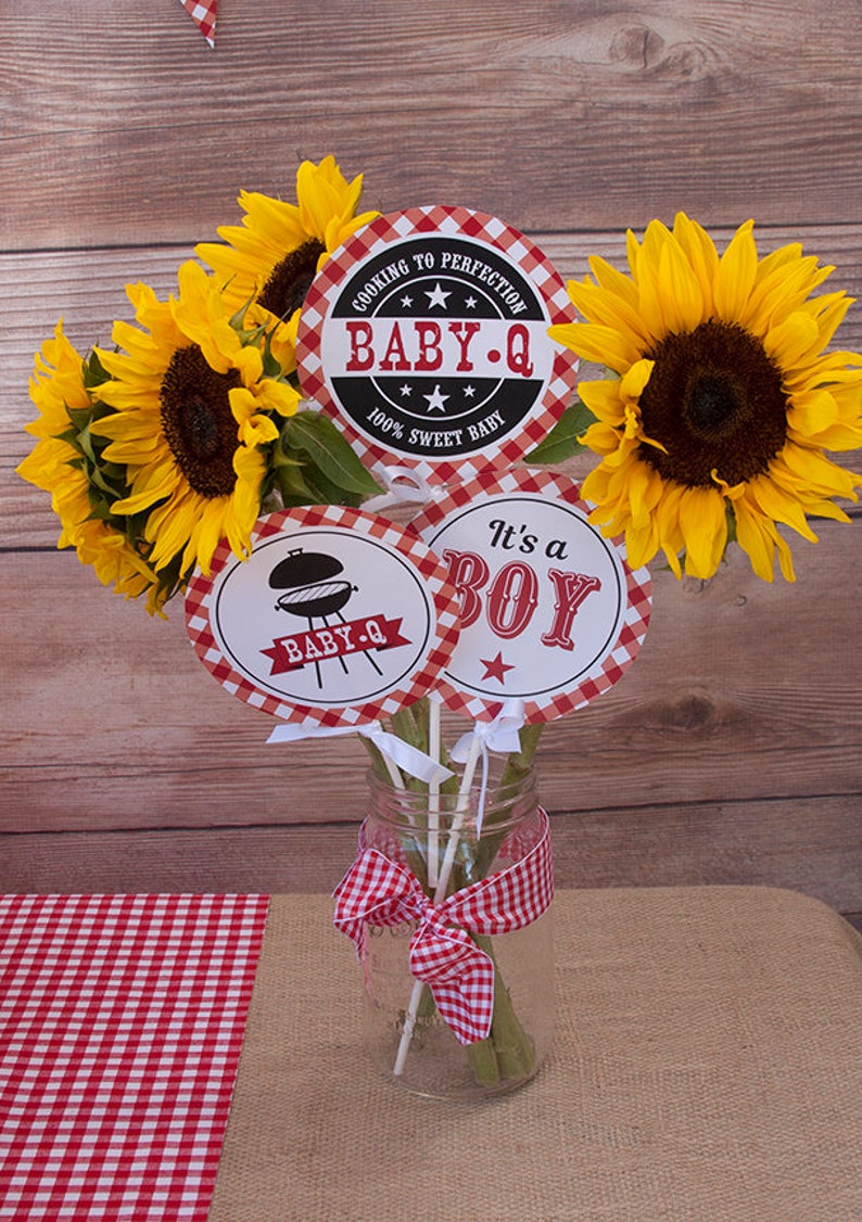 Może przedstawiać: Centralny element dekoracyjny na baby shower z motywem grillowania. Trzy okrągłe papierowe tabliczki z czerwono-białym wzorem w kratkę. Tabliczki przedstawiają napisy "Cooking to Perfection Baby-Q 100% Sweet Baby", "Baby-Q" z obrazkiem grilla oraz "It's a Boy" z czerwoną gwiazdą. Tabliczki są przymocowane do drewnianych szaszłyk&oacute;w i umieszczone w przezroczystym szklanym słoiku z czerwoną i białą wstążką w kratkę. Słoik jest otoczony słonecznikami.