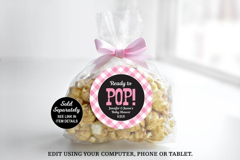 May include: A clear plastic bag filled with popcorn, tied with a pink ribbon. A round sticker with a pink and white gingham pattern and black text reads "Ready to POP! Jennifer & Jason's Baby Shower 8.12.21". A smaller black circle sticker reads "Sold Separately SEE LINK IN ITEM DETAILS".