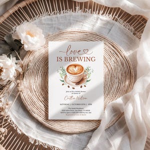 K&ouml;nnte beinhalten: Eine Einladung zur Brautparty mit dem Text "love is brewing" und einer Kaffeetassen-Illustration. Die Einladung befindet sich auf einer wei&szlig;en Karte, umgeben von einem gewebten Platzset, wei&szlig;en Blumen und einem Leinentuch.