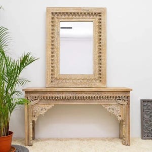 May include: A light-colored wooden console table with a matching framed mirror. The table has carved details along the top and arched supports. The mirror is rectangular with an ornate frame. A potted plant is on the left.