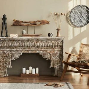 May include: A living room interior with a carved wooden console table and various decorative elements. A black sculpture, dried flowers in a vase, and a woven chair with a blanket are visible. The room has a neutral colour scheme.