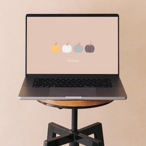 Op de afbeelding: Een laptop met een scherm met een lichtbruine achtergrond met vier kleine, eenvoudige pompoenillustraties in geel, wit, saliegroen en donkerbruin, met het woord Autumn in een cursief wit lettertype eronder in het midden geplaatst.