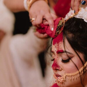 May include: A close-up of a woman's face during a traditional ceremony. Her forehead and nose are adorned with red powder. She wears gold jewelry, including a nose ring and earrings. A hand holds a red cloth above her head.