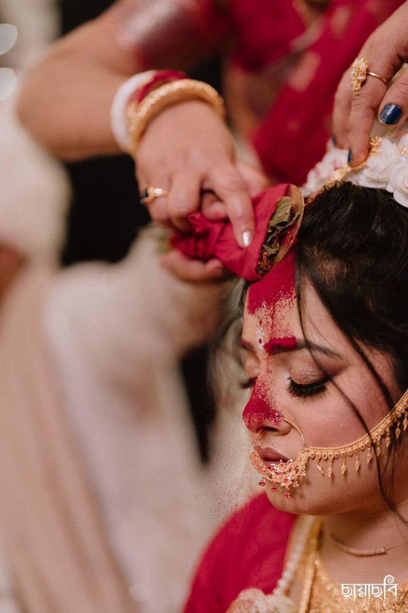 May include: A close-up of a woman's face during a traditional ceremony. Her forehead and nose are adorned with red powder. She wears gold jewelry, including a nose ring and earrings. A hand holds a red cloth above her head.
