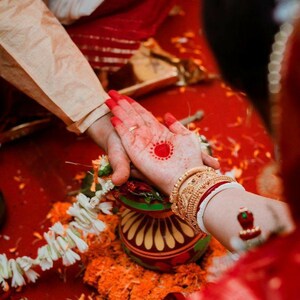 May include: Ceremonial hands clasped during a traditional Indian wedding. The bride's hand is adorned with henna and gold bangles. A red dot is painted on the palm. The background is a vibrant red.