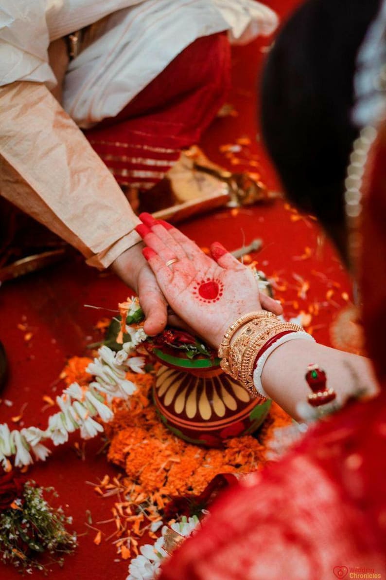 May include: Ceremonial hands clasped during a traditional Indian wedding. The bride's hand is adorned with henna and gold bangles. A red dot is painted on the palm. The background is a vibrant red.