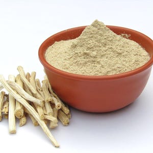 Puede incluir: Un cuenco de terracota lleno de polvo marr&oacute;n claro, probablemente una especia o suplemento, se encuentra junto a un mont&oacute;n de tallos de plantas beige y secos. Los elementos est&aacute;n sobre un fondo blanco.