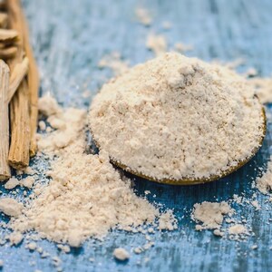 Puede incluir: Primer plano de polvo de ashwagandha y ra&iacute;ces de ashwagandha secas sobre una superficie de madera azul. El polvo es de color beige claro y est&aacute; en un plato peque&ntilde;o, redondo y dorado. Las ra&iacute;ces son de color marr&oacute;n claro y tienen una textura rugosa.