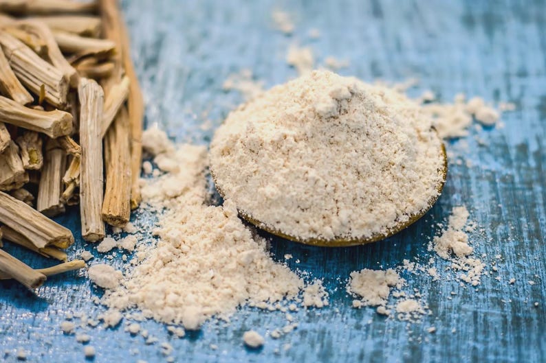 Puede incluir: Primer plano de polvo de ashwagandha y ra&iacute;ces de ashwagandha secas sobre una superficie de madera azul. El polvo es de color beige claro y est&aacute; en un plato peque&ntilde;o, redondo y dorado. Las ra&iacute;ces son de color marr&oacute;n claro y tienen una textura rugosa.