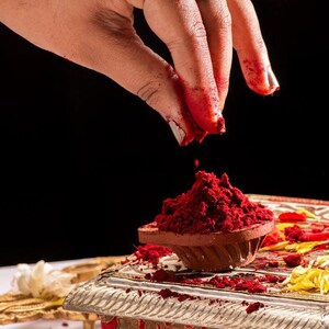 May include: A hand with red powder on the fingers sprinkles more red powder into a small, terracotta bowl. The bowl sits on a decorative, silver-colored stand with red and gold accents. A large ring with a dark stone adorns the hand.