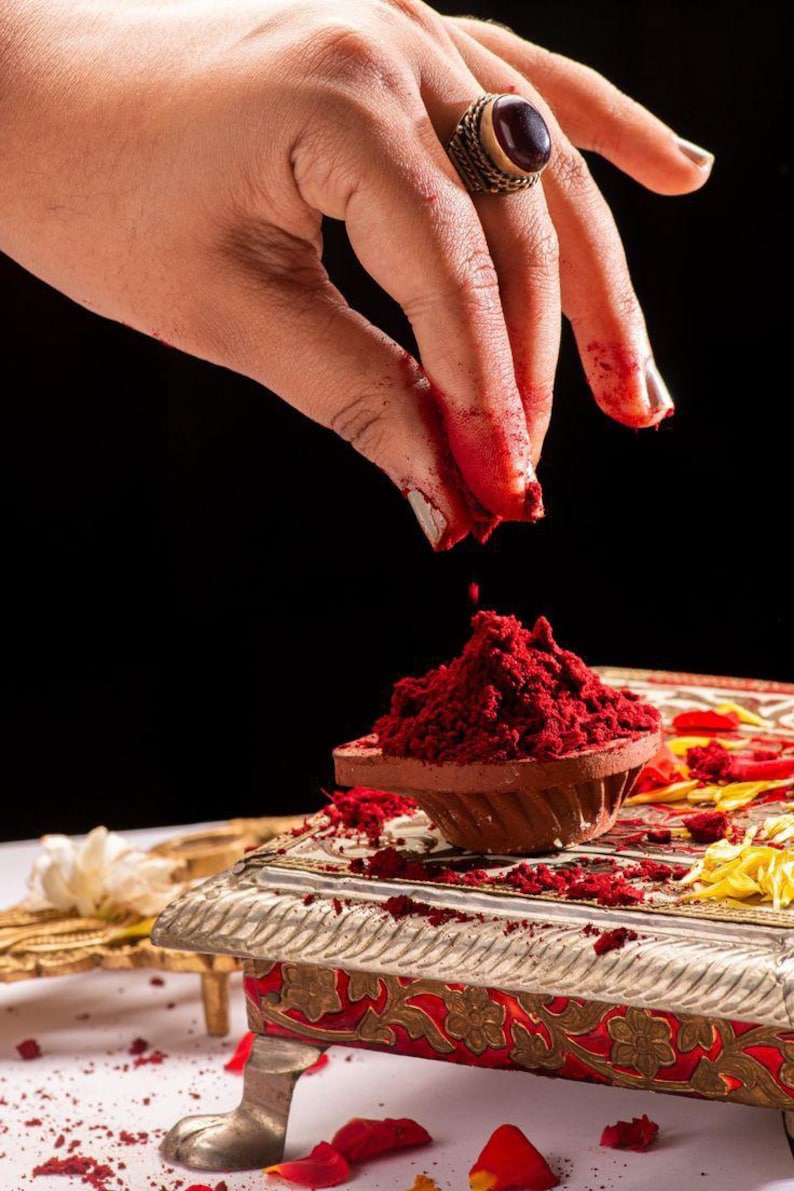 May include: A hand with red powder on the fingers sprinkles more red powder into a small, terracotta bowl. The bowl sits on a decorative, silver-colored stand with red and gold accents. A large ring with a dark stone adorns the hand.