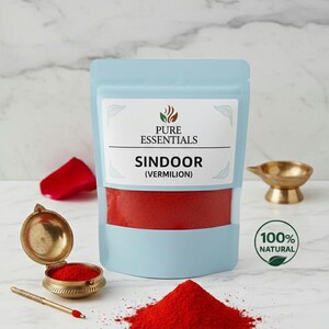 May include: A light blue pouch labeled "PURE ESSENTIALS" containing red sindoor (vermilion) powder. A small pile of the red powder sits in front of the pouch. A small, open, gold-colored container with a red powder is to the left.