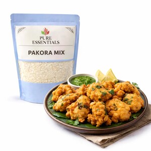 May include: A bag of Pure Essentials Pakora Mix next to a plate of golden-brown pakoras, garnished with green herbs. A small bowl of green chutney and lemon wedges complete the presentation. The bag has a light blue background.