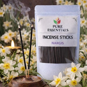Könnte beinhalten: Eine Packung Pure Essentials Nargis Räucherstäbchen wird gezeigt, mit einem transparenten Fenster, das die dunklen Stäbchen zeigt. Zwei Räucherstäbchen brennen in einem Holzhalter und geben Rauch ab. Der Hintergrund zeigt weiße und gelbe Blumen und eine brennende Kerze.