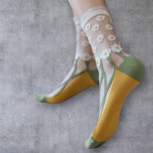 Pode incluir: Meias transparentes com padrão floral. As meias apresentam flores brancas com centros amarelos, caules verdes e detalhes amarelos e verdes no calcanhar e na ponta. A parte superior da meia é branca.