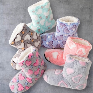 Puede incluir: Una colección de seis botas de estar por casa de felpa hasta el tobillo con un suave forro polar blanco. Cada par muestra un patrón repetido de corazones en varias combinaciones de colores, incluyendo verde menta, lavanda, rosa coral y gris.