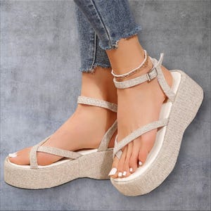 May include: A pair of beige platform sandals featuring a textured fabric finish, a thick sole, and thin straps that wrap around the ankle and across the top of the foot. The sandals have a white footbed and a secure buckle closure at the ankle.