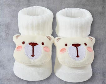 May include: A pair of white baby socks with a cream-colored bear face design on the front. The bear has brown embroidered details for the eyes and nose, and pink cheeks. The socks have a ribbed cuff.