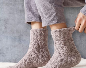May include: A pair of soft, fluffy, pale purple socks. The socks have a thick, textured appearance with a ribbed cuff. The person is wearing matching pale purple trousers. The socks are on a white surface.