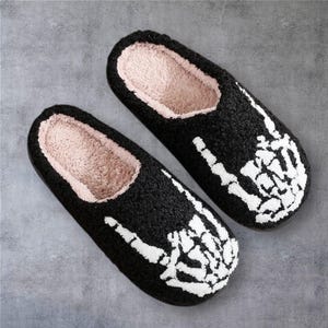 May include: A pair of black slippers with a white skeleton hand graphic on the top. The slippers feature a textured black exterior and a soft, light pink plush interior lining. The design shows a skeletal hand making a rock and roll gesture.