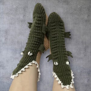 Könnte beinhalten: Ein Paar olivgrüne Häkelsocken, die wie Krokodile gestaltet sind. Die Socken verfügen über strukturierte Rippen auf der Oberseite, kleine Seitenflossen, weiße gehäkelte Zähne am Knöchelbereich sowie weiß-schwarze Augen auf dem Fußrücken.