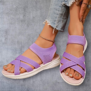 Op de afbeelding: Een paar lavendelkleurige gebreide sandalen met een kruislings bandontwerp over de voorvoet, een stevige enkelband en een witte, gedempte zool. Het lichtgewicht stoffen bovenwerk zorgt voor een flexibele pasvorm en een platte basis.