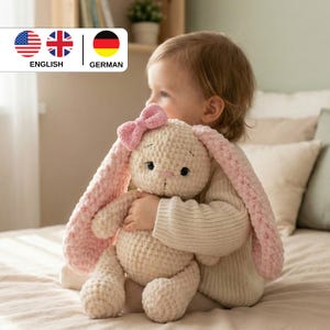 May include: A handmade, cream-colored crochet bunny with long, pink ears and a pink bow. The stuffed animal is being held by a child. The bunny has black eyes and a small pink nose. The background is a soft, neutral color.