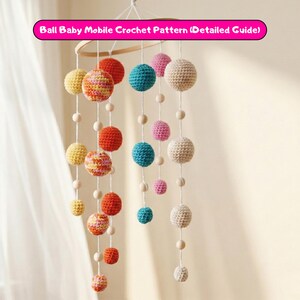 May include: A handmade baby mobile featuring crochet balls in various colors, including yellow, orange, pink, blue, and cream. The balls are suspended from a wooden hoop with wooden beads. The text "Ball Baby Mobile Crochet Pattern (Detailed Guide)" is visible.