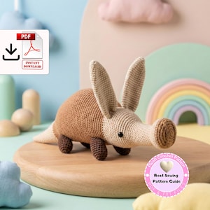 Può includere: Un peluche di oritteropo all'uncinetto nei toni del marrone e beige, con un lungo muso e grandi orecchie. Il giocattolo è su una superficie di legno, con un'icona di download PDF e un distintivo rosa con la scritta "Best Sewing Pattern Guide".