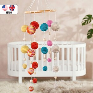 May include: A colorful baby mobile with crochet spheres in various sizes and colors, including yellow, red, pink, blue, and cream. The spheres are suspended from a wooden hoop with wooden beads. The mobile hangs in front of a white crib.