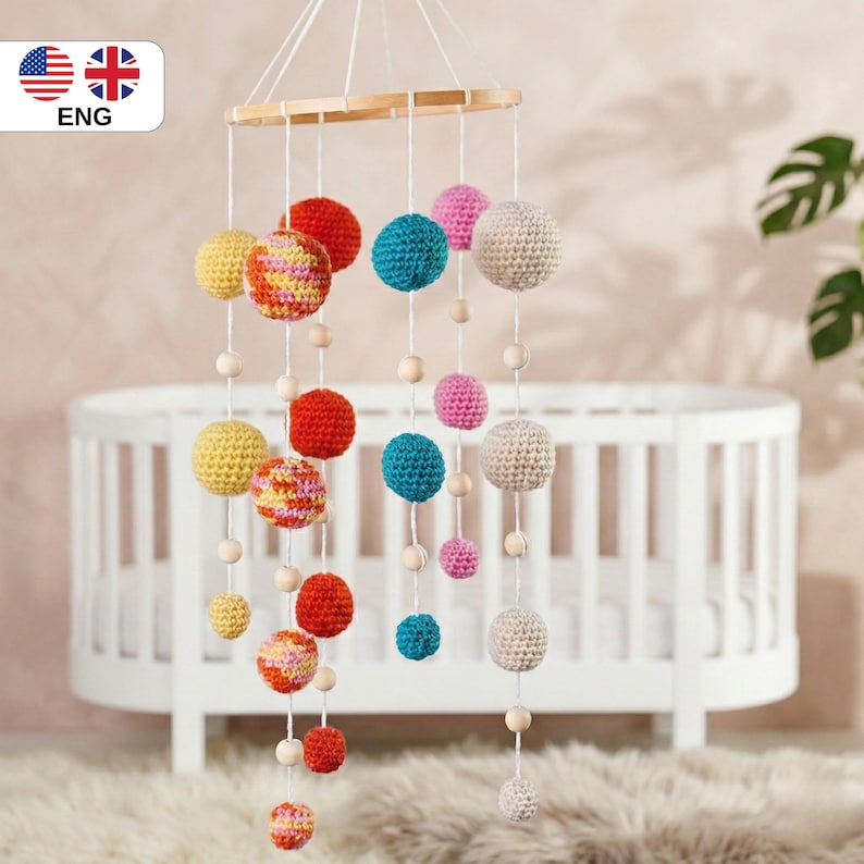 May include: A colorful baby mobile with crochet spheres in various sizes and colors, including yellow, red, pink, blue, and cream. The spheres are suspended from a wooden hoop with wooden beads. The mobile hangs in front of a white crib.