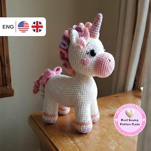 May include: A handmade white crochet unicorn with a pink muzzle, cheeks, and hooves. The unicorn has a multicolored mane and tail, and a spiraled horn. The plush toy is standing on a wooden surface. The image includes text that says "Plush Crochet Pattern" and "Best Sewing Pattern Guide".