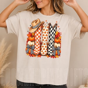 Puede incluir: Una camiseta beige con un gráfico de tres botas vaqueras, una con patrón de cuadros naranjas, otra con estampado de calabazas y otra con cuadros negros, rodeadas de hojas de otoño, calabazas y un sombrero de vaquero.