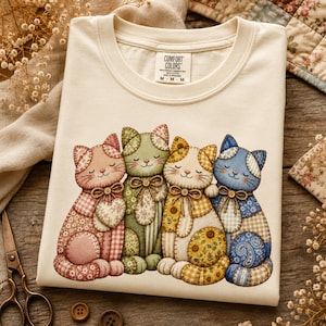 May include: A cream-colored crew neck t-shirt featuring a graphic of four cats sitting side-by-side. Each cat is designed with various patchwork patterns in shades of pink, green, yellow, and blue, and each wears a small brown bow around its neck.