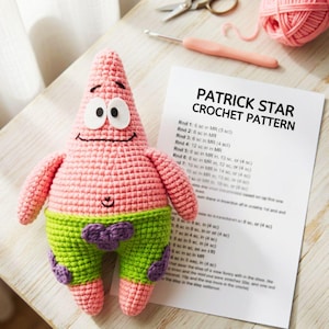 Puede incluir: Un muñeco de Patrick Estrella hecho a mano en ganchillo rosa con ojos blancos, boca negra y pantalones cortos verdes con patrones de flores moradas. Junto al muñeco hay una hoja blanca con el texto impreso PATRICK STAR CROCHET PATTERN.
