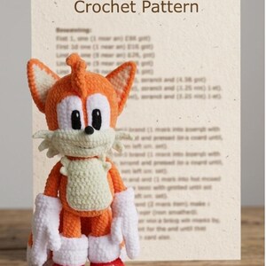 May include: A crocheted plush toy of a fox character with orange fur, a cream-colored chest and muzzle, large white gloves, and red and white shoes. Behind the toy is a document titled Copter-tailed Fox Crochet Pattern with blurred text.