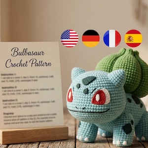 May include: A light blue and green crochet creature with red eyes and a bulbous plant on its back, positioned next to a rectangular sign in a wooden stand that reads Bulbasaur Crochet Pattern and includes written instructions.