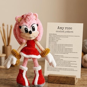 May include: A crocheted Amy Rose doll with pink fur, a red dress, white gloves, and red boots with white trim. Next to the doll is a rectangular card on a wooden stand with the printed text Amy rose crochet pattern and instructions.
