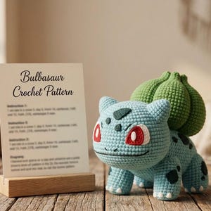 Könnte beinhalten: Eine hellblaue und grüne Häkelfigur mit roten Augen und einer knolligen Pflanze auf dem Rücken, neben einer weißen Karte in einem Holzständer, die mit Bulbasaur Crochet Pattern beschriftet ist und Anleitungen für das Projekt enthält.