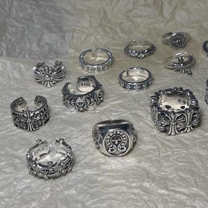 Op de afbeelding: Een verzameling zilveren ringen met diverse ontwerpen, waaronder kruisen, bloemmotieven en geometrische vormen. Sommige ringen hebben gegraveerde details en data. De ringen worden gepresenteerd op een getextureerd, gebroken witte ondergrond.