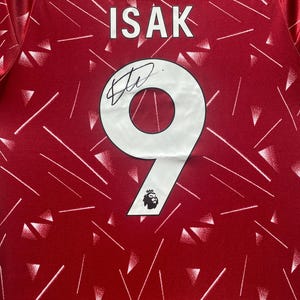 May include: A red sports jersey featuring a white geometric pattern, the name ISAK printed in white, and a large white number 9 with a black signature across it. A small white emblem with the number 97 is visible at the top centre.