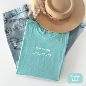 以下が含まれることがあります： 「seas the day」の文字と波のグラフィックが入った水色のTシャツ、薄い色のジーンズ、ベージュの帽子、ネックレスを組み合わせたカジュアルなファッションのフラットレイ。Tシャツの色は「Chalky Mint」です。