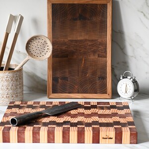 Peut inclure: Scène de cuisine avec une planche à découper en bois et un couteau, une planche en bois encadrée et un petit réveil argenté. Un porte-ustensiles en céramique contient un fouet, des spatules et une cuillère ajourée.