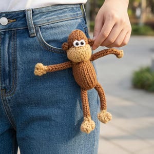 Könnte beinhalten: Ein braunes und beigefarbenes Strickaffen-Spielzeug mit ausgestreckten Armen und Beinen. Der Affe hat weiße Augen mit schwarzen Pupillen und wird in der Nähe einer blauen Jeans-Tasche gehalten.